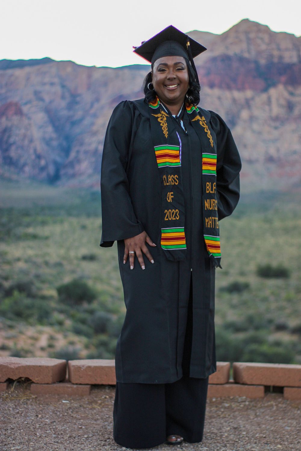 Nursing graduate in cap and gown with Class of 2023 kente stole at Red Rock Canyon.