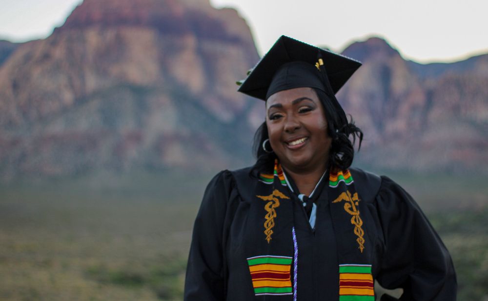 Smiling woman in graduation cap and gown with colorful stole poses before red rock mountains.