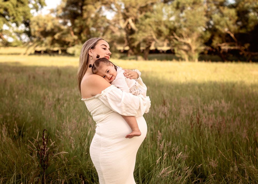 A pregnant woman in a flowing white dress stands in a golden field at sunset, cradling her baby and baby bump.