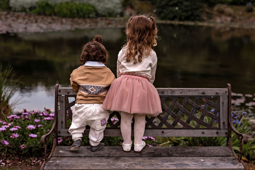 Two children sitting together on a wooden bench by a serene pond surrounded by nature and flowers.