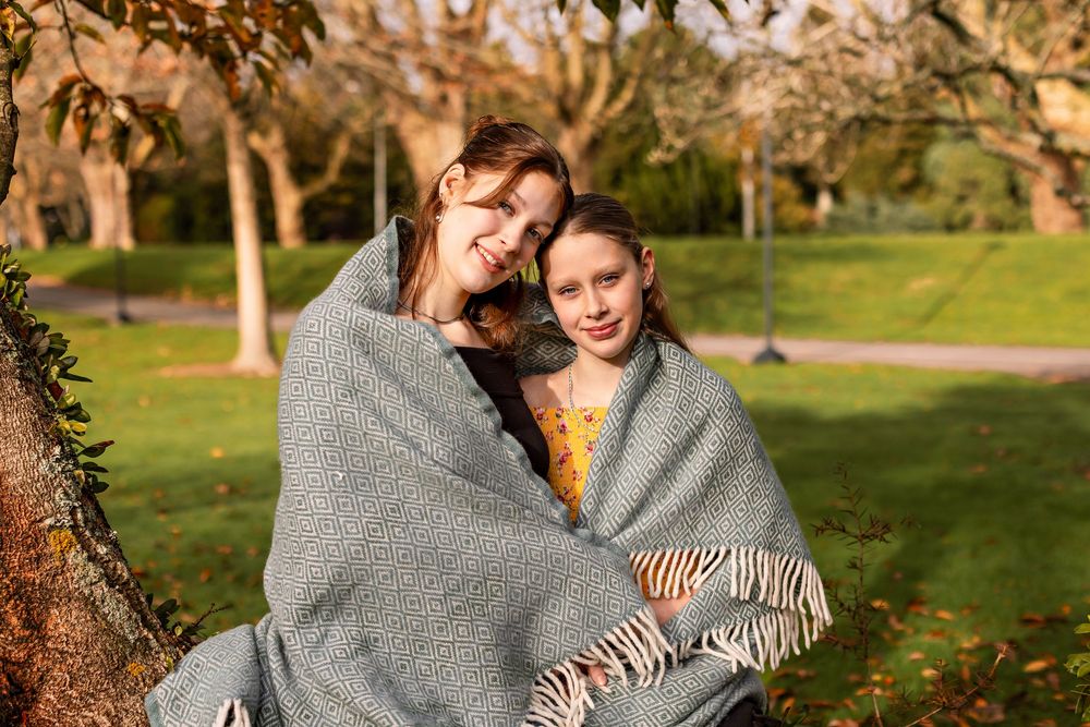 A couple of sisters wrapped in a cozy grey blanket shares tender moments in a sunny autumn park setting. Bledisloe Park Palmerston North