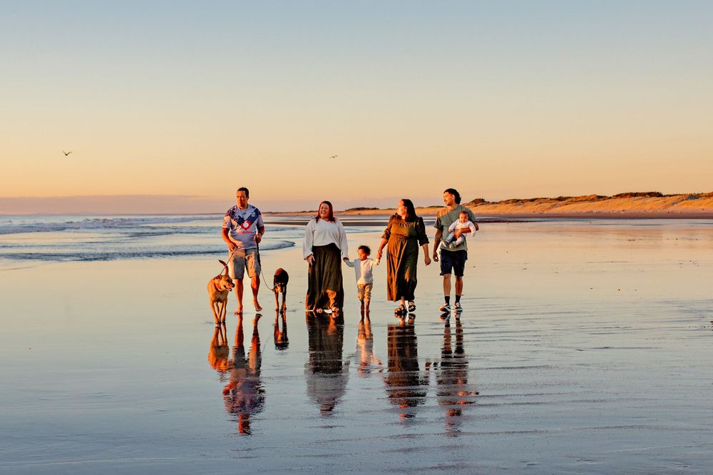 A family stands on the reflective sand of Foxton Beach at sunset. The sky glows with warm golden and orange hues.