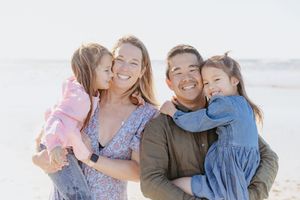 A family of four shares joyful embraces while posing together on a bright, sunny beach.