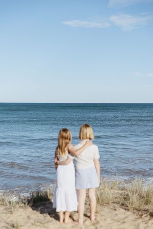 Two children in white dresses stand together looking out at the ocean from a sandy beach on a sunny day.