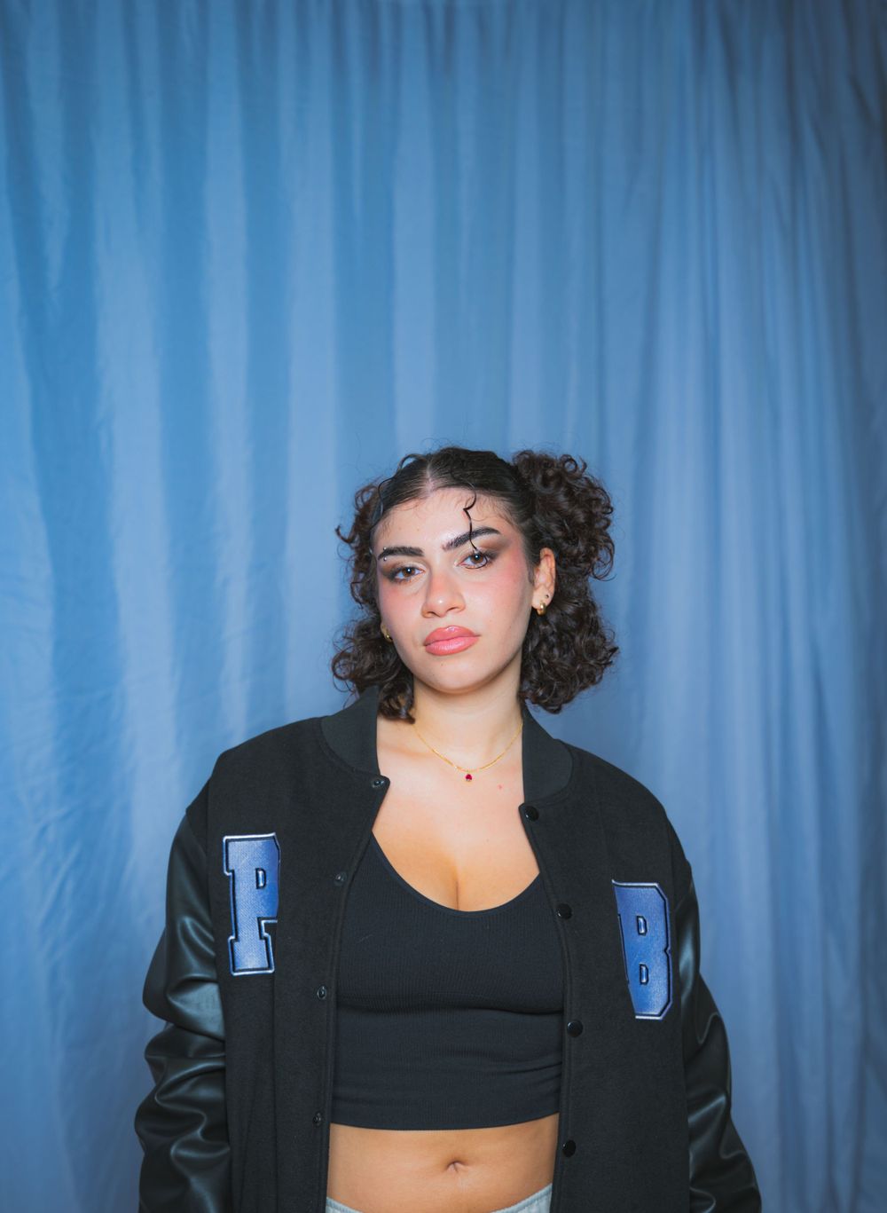 Fashion portrait with varsity jacket and black crop top against blue curtain background.