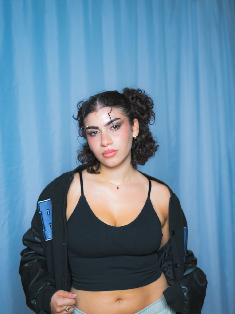 Portrait against blue curtain backdrop featuring black crop top and letterman jacket styling.