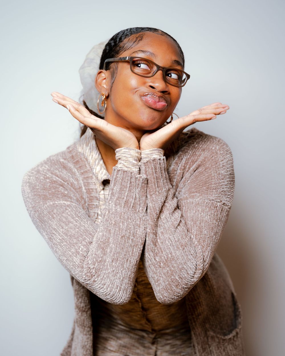 Portrait of person in glasses wearing cozy sweater making playful expression with hands under chin.