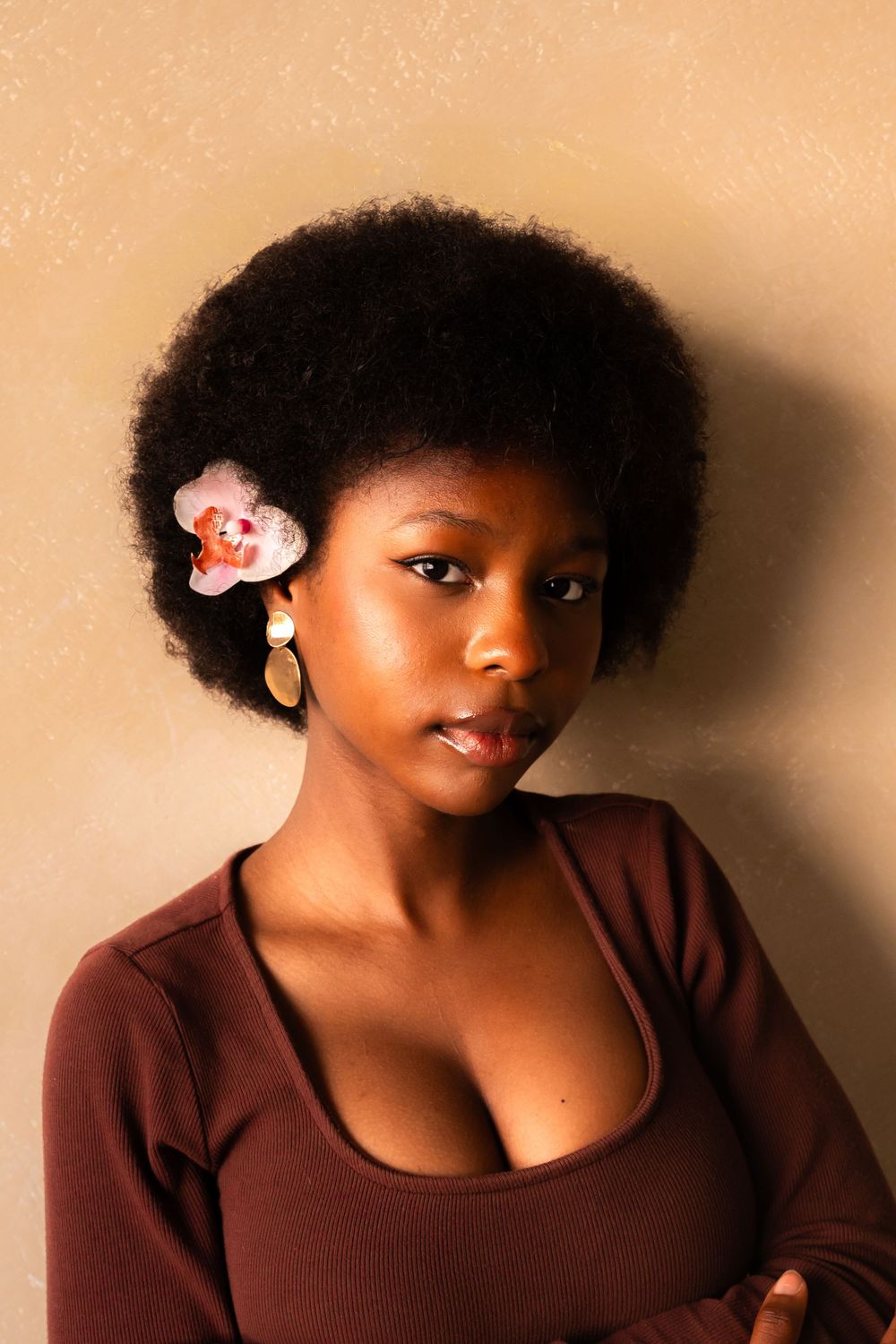 Portrait against beige wall showing natural afro hairstyle with small flower accessory and brown top.