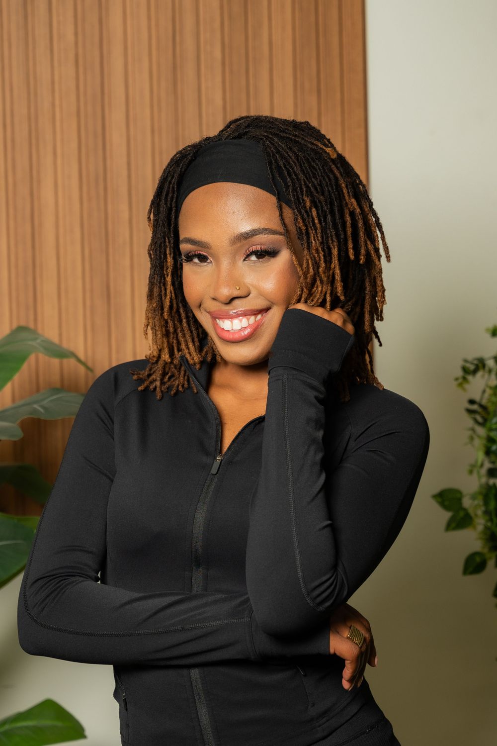 Professional portrait of someone wearing a black zip-up jacket and styled locs against a warm-toned background.