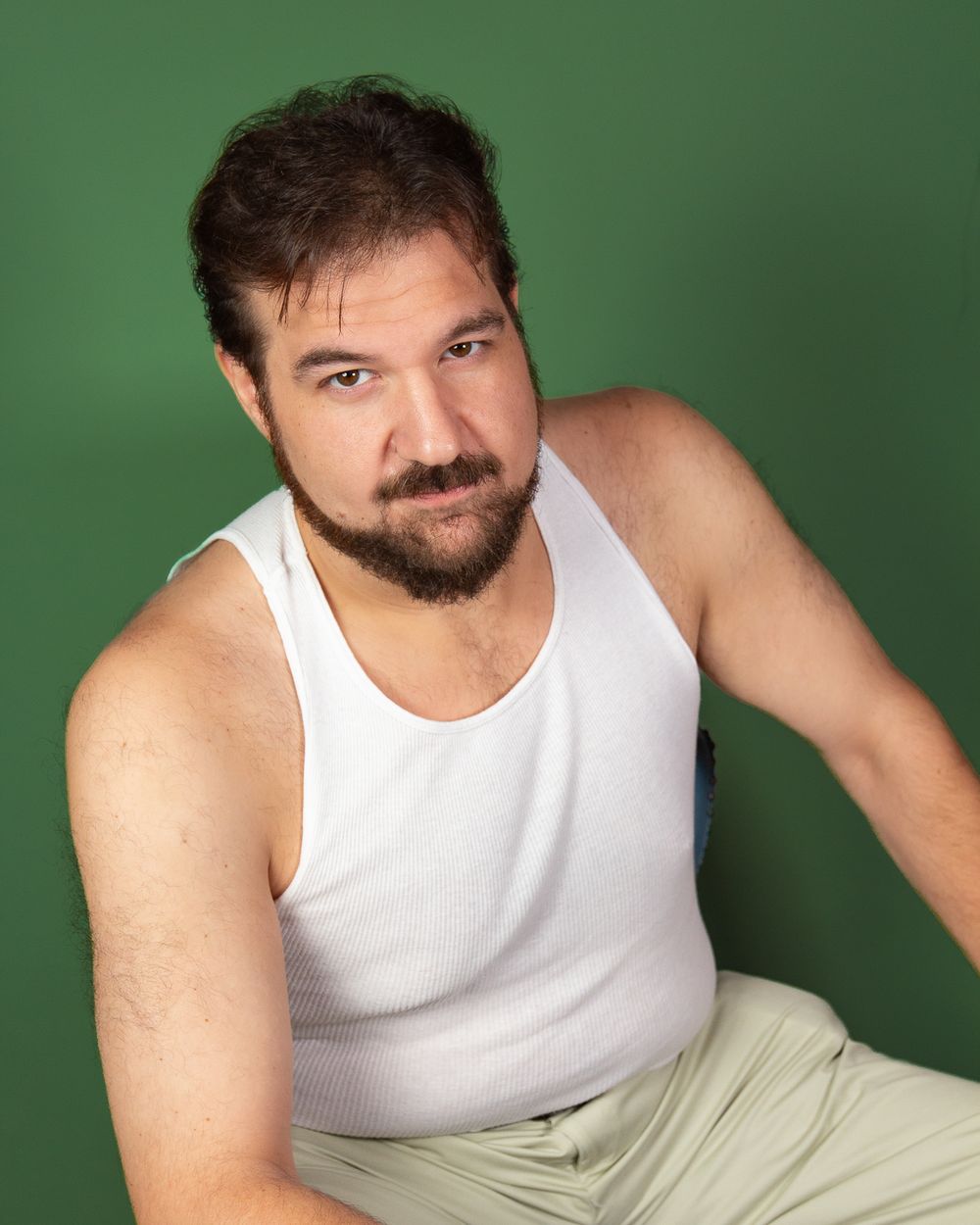 Studio portrait of a bearded person in a white tank top against a green background.