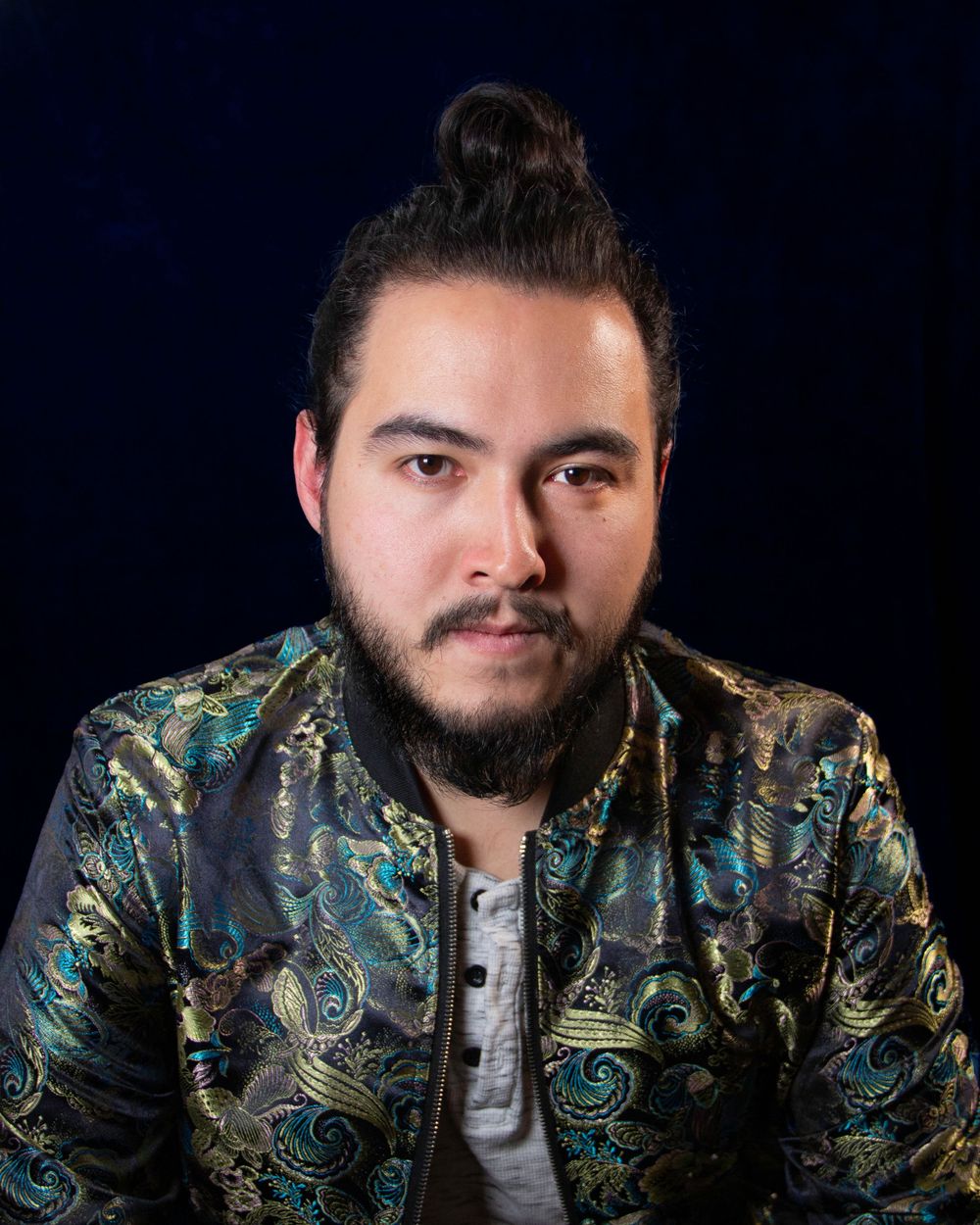 Portrait of person in ornate patterned jacket against dark background with hair in top knot style.