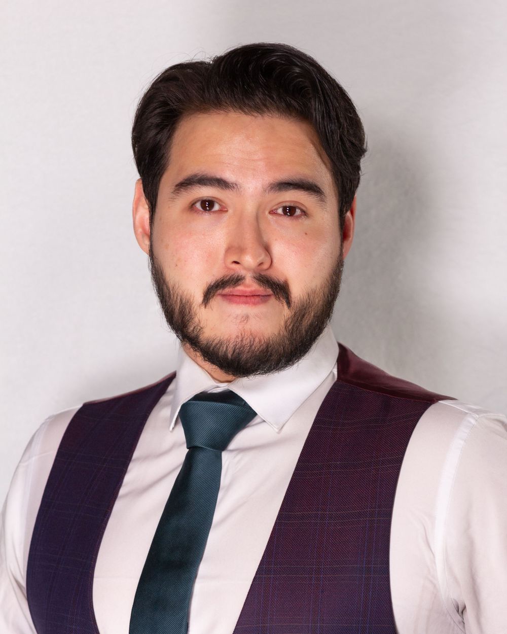 Professional headshot of person wearing white shirt, blue tie and burgundy vest against light background.