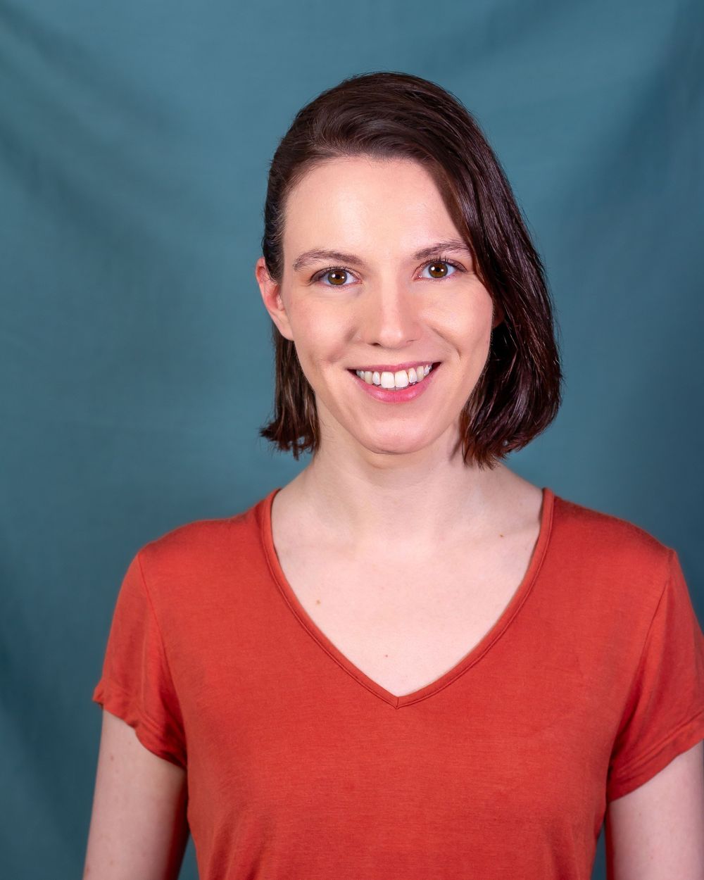 Professional headshot of someone in a red v-neck shirt against a teal backdrop.