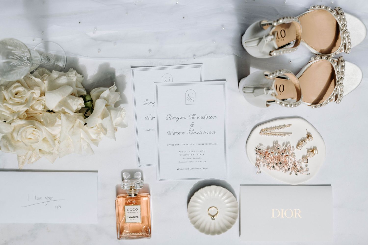Elegant wedding details flatlay with perfume, invitation, shoes and white flowers on white surface.