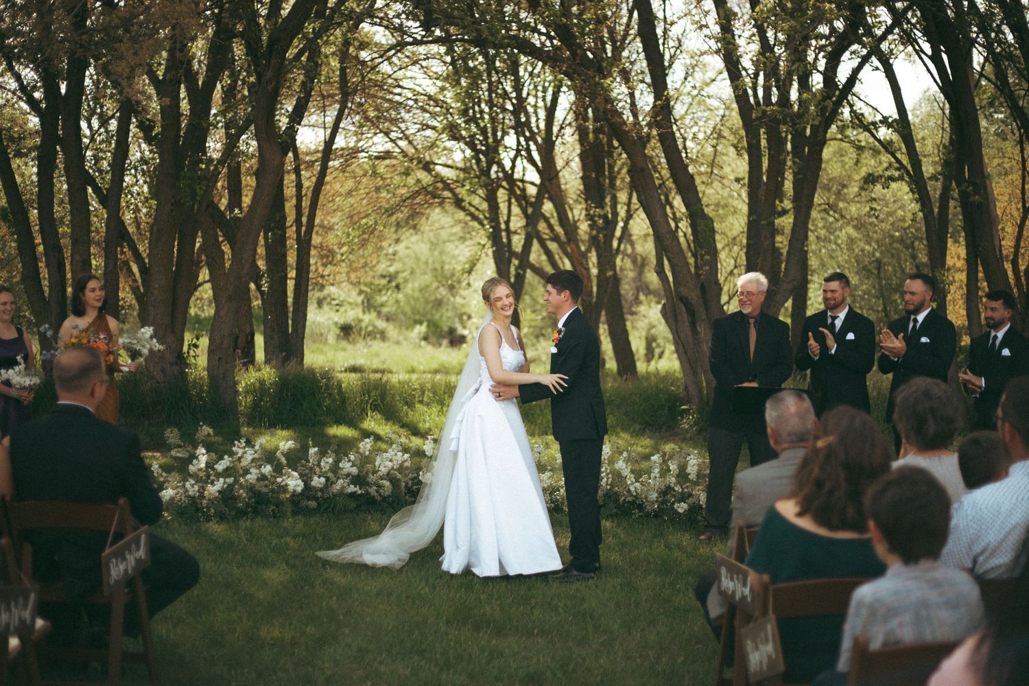 A wedding ceremony takes place in a wooded outdoor setting with guests seated on white chairs.