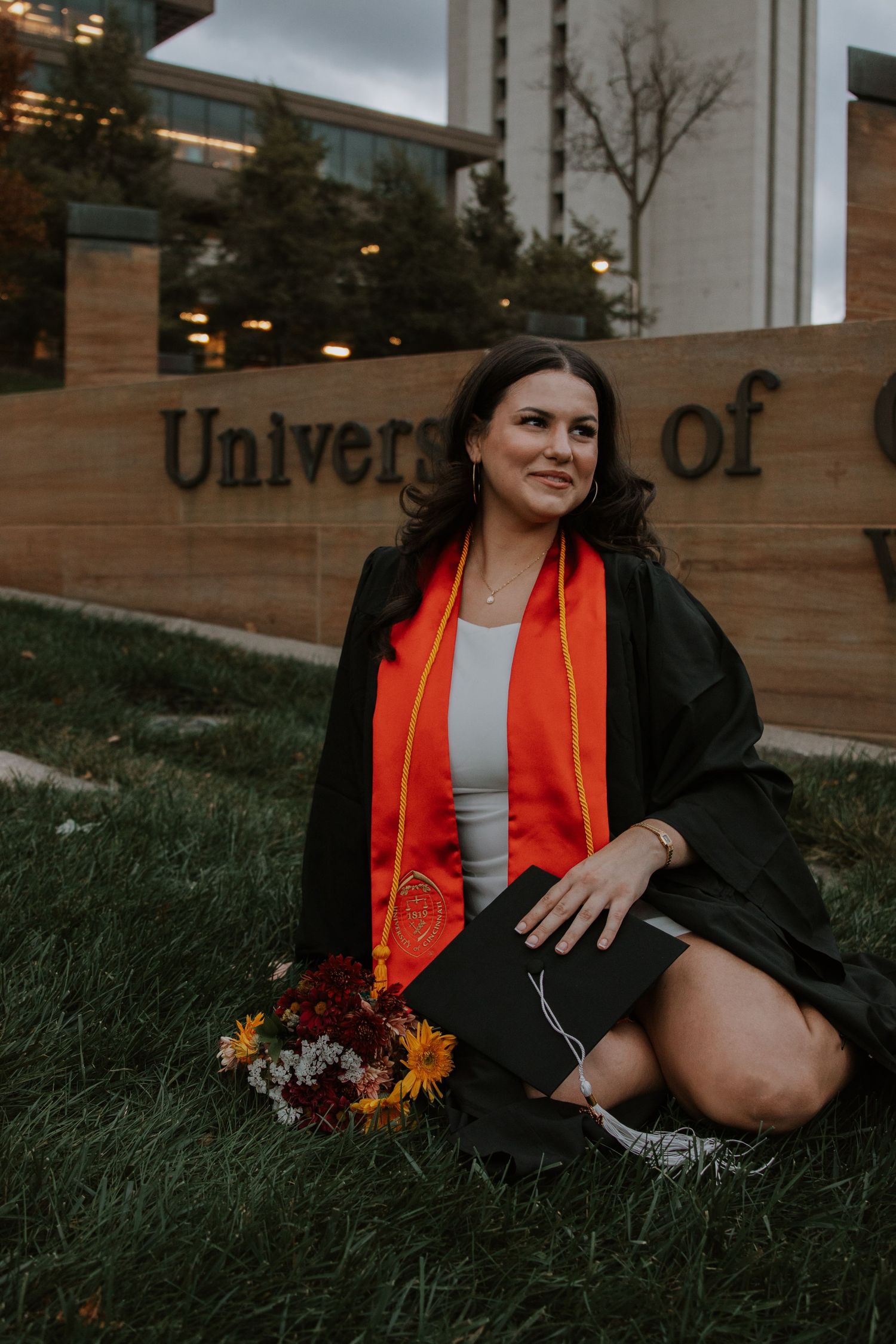 Graduate in cap and gown posing with flowers at university sign.