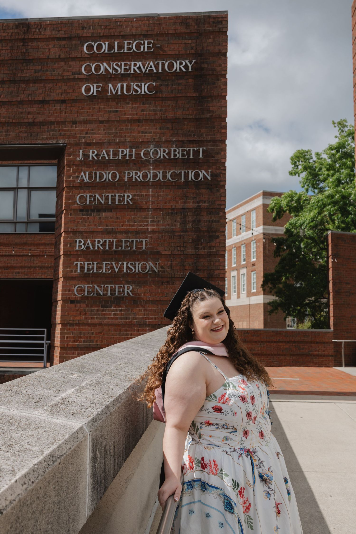 Graduate in floral dress and cap poses proudly in front of College Conservatory of Music building.