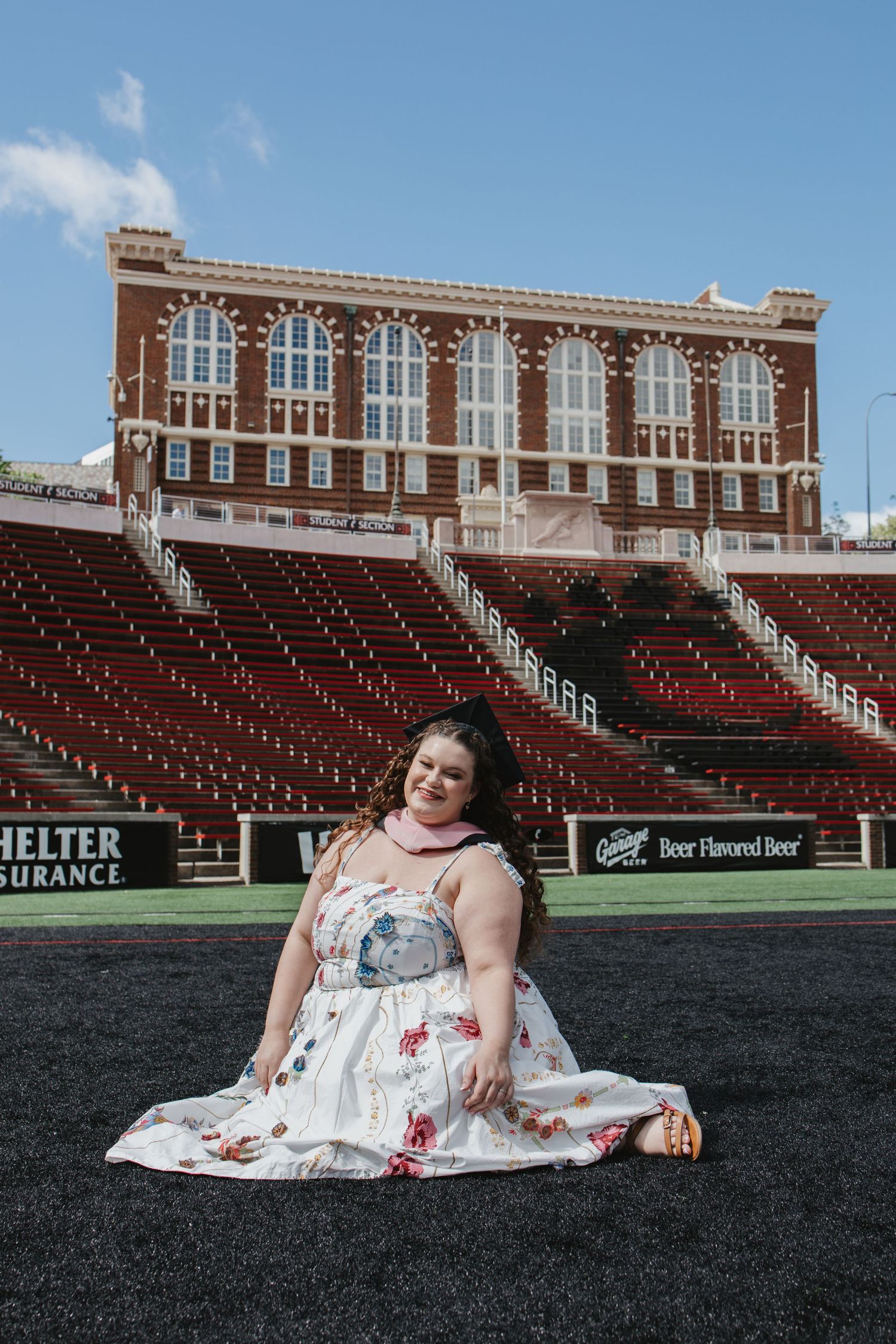 Woman in floral dress sitting on football field with historic brick stadium stands in background.