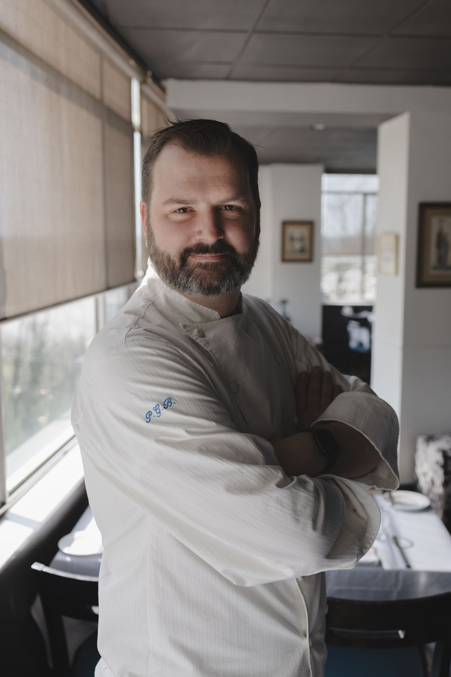 Professional chef in white coat stands with arms crossed, smiling confidently in an elegant restaurant setting.