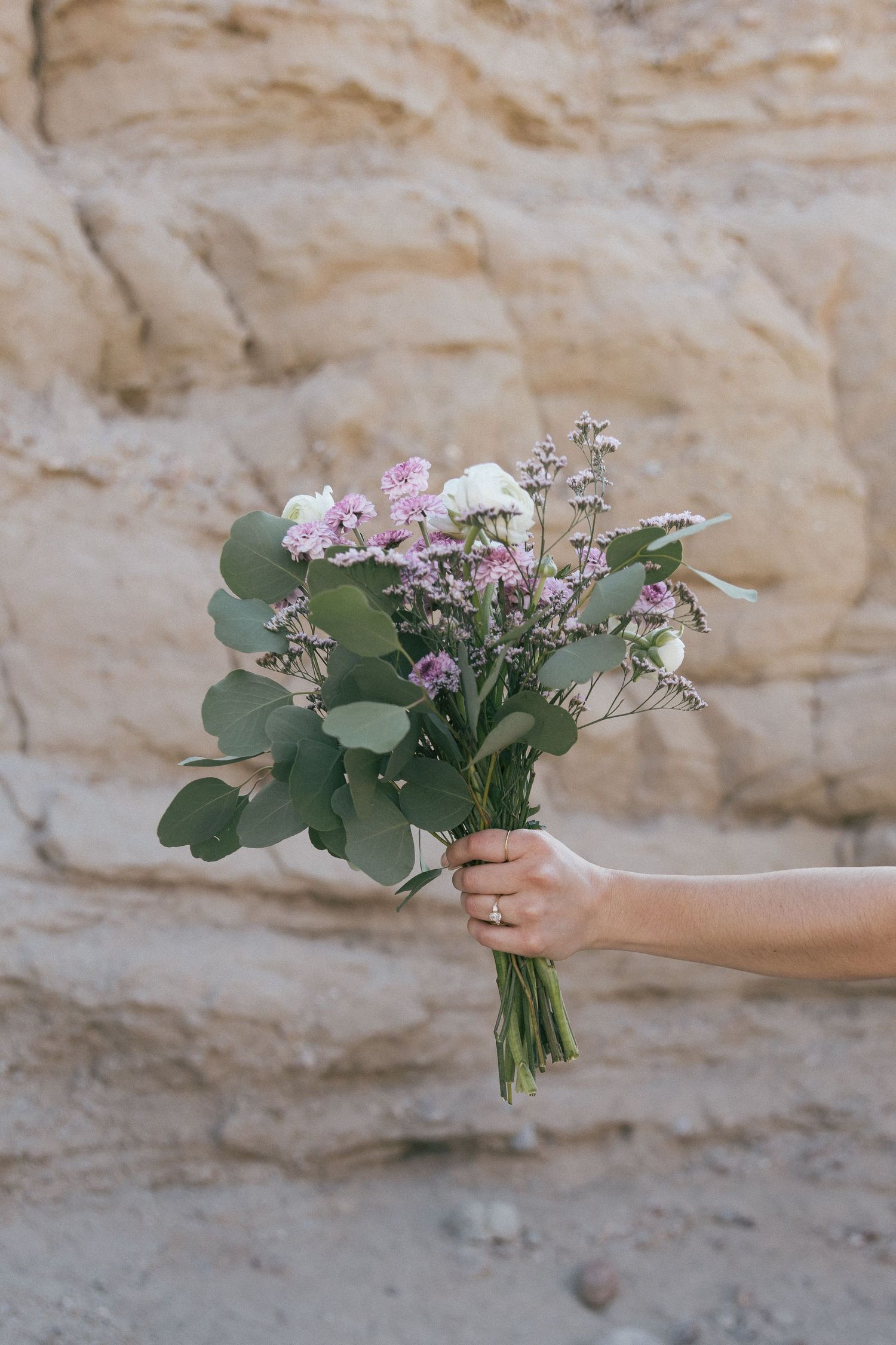 Hand holding delicate bouquet of purple flowers and eucalyptus against limestone wall.