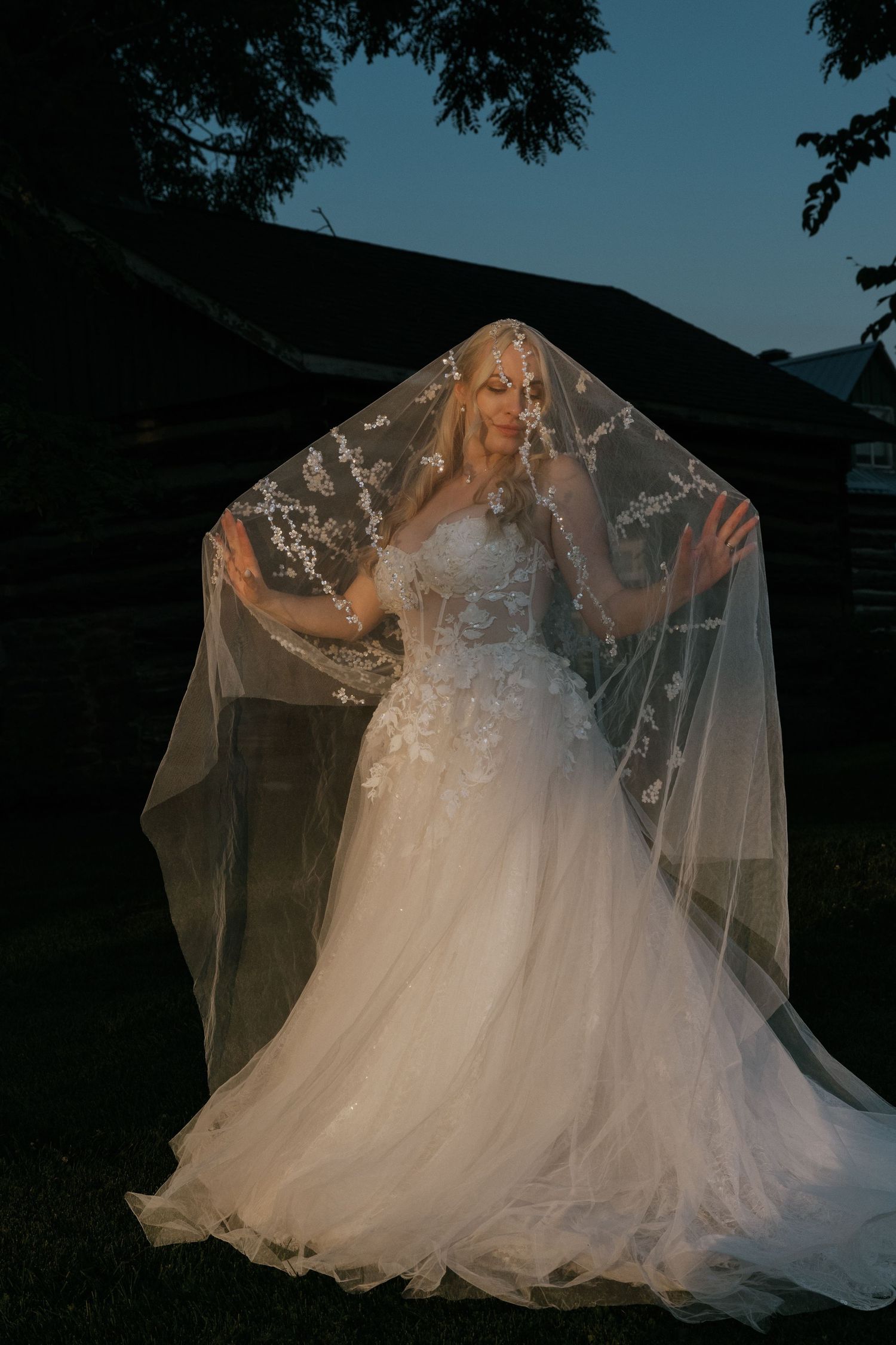 Elegant bride in white lace wedding gown poses in garden setting with small floral bouquet.