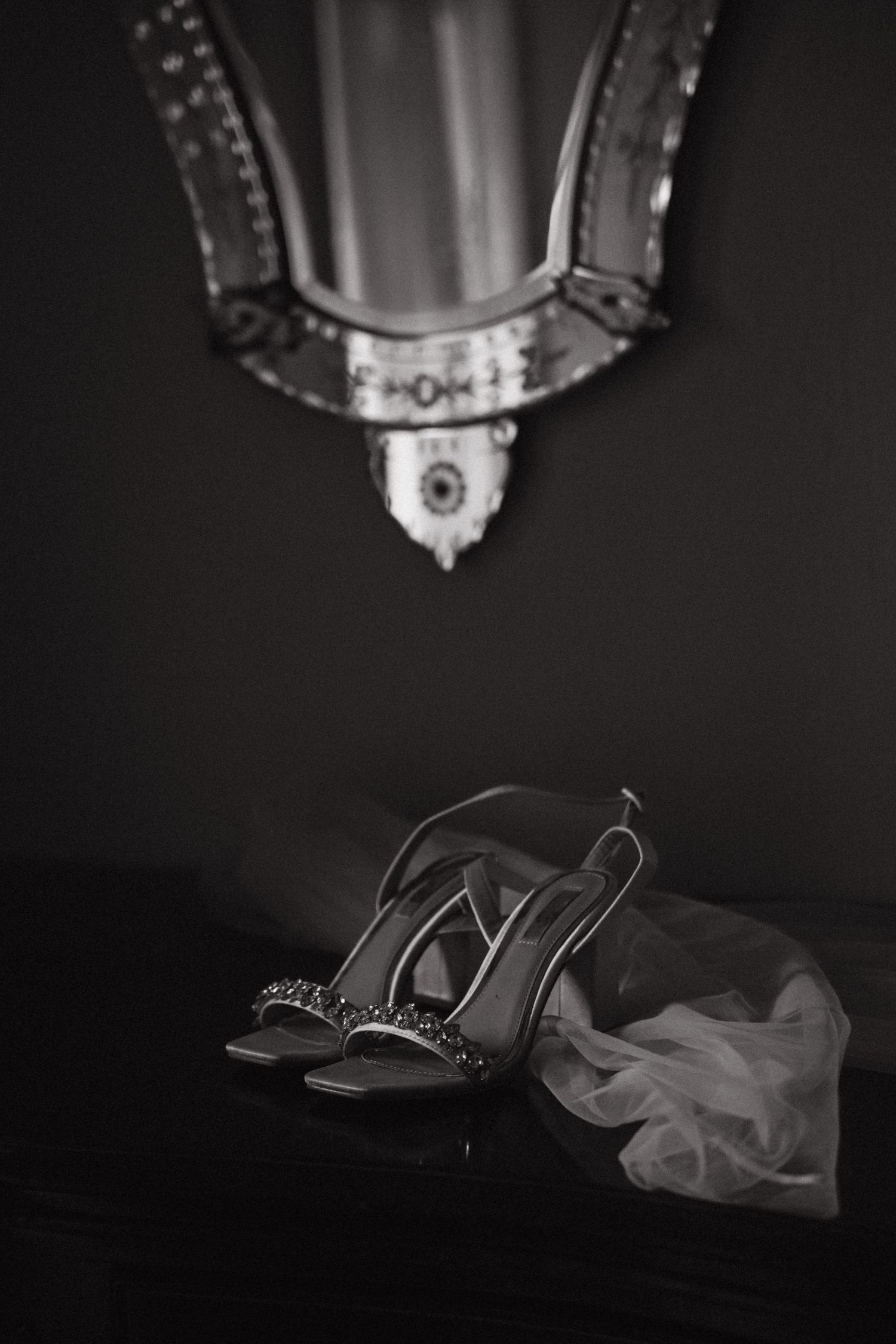 Black and white still life of elegant bridal shoes with ornate mirror above.