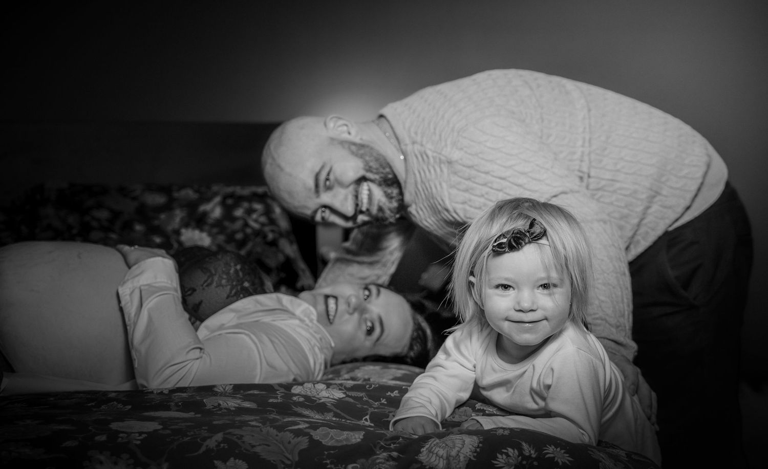 Maman allongée sur le lit, papa souriant derrière leur fille, en noir et blanc à Ange-Gardiens, Québec.