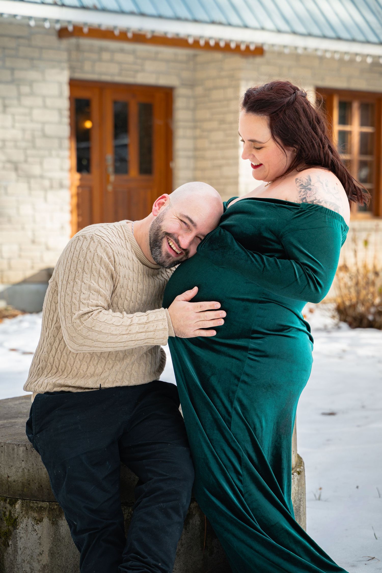 Papa souriant la tête sur le ventre de la maman enceinte en robe verte, dehors dans la neige à Ange-Gardiens, Québec.