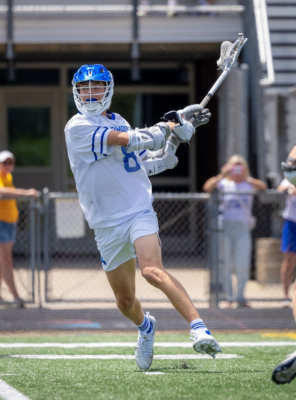 Lacrosse player in white and blue uniform mid-swing with stick on turf field during sunny outdoor game.