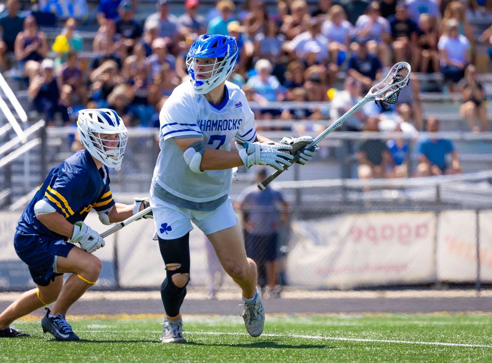 Two lacrosse players compete during a sunny outdoor game, one in blue Shamrocks uniform cradling the ball.