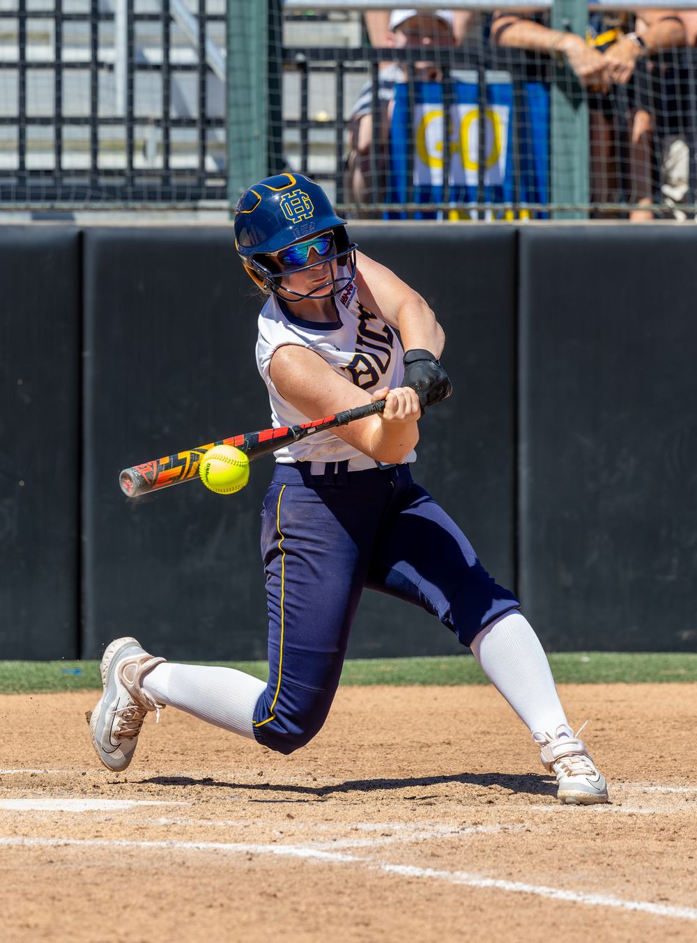 Female softball player in blue uniform making contact with a pitch during a sunny outdoor game.
