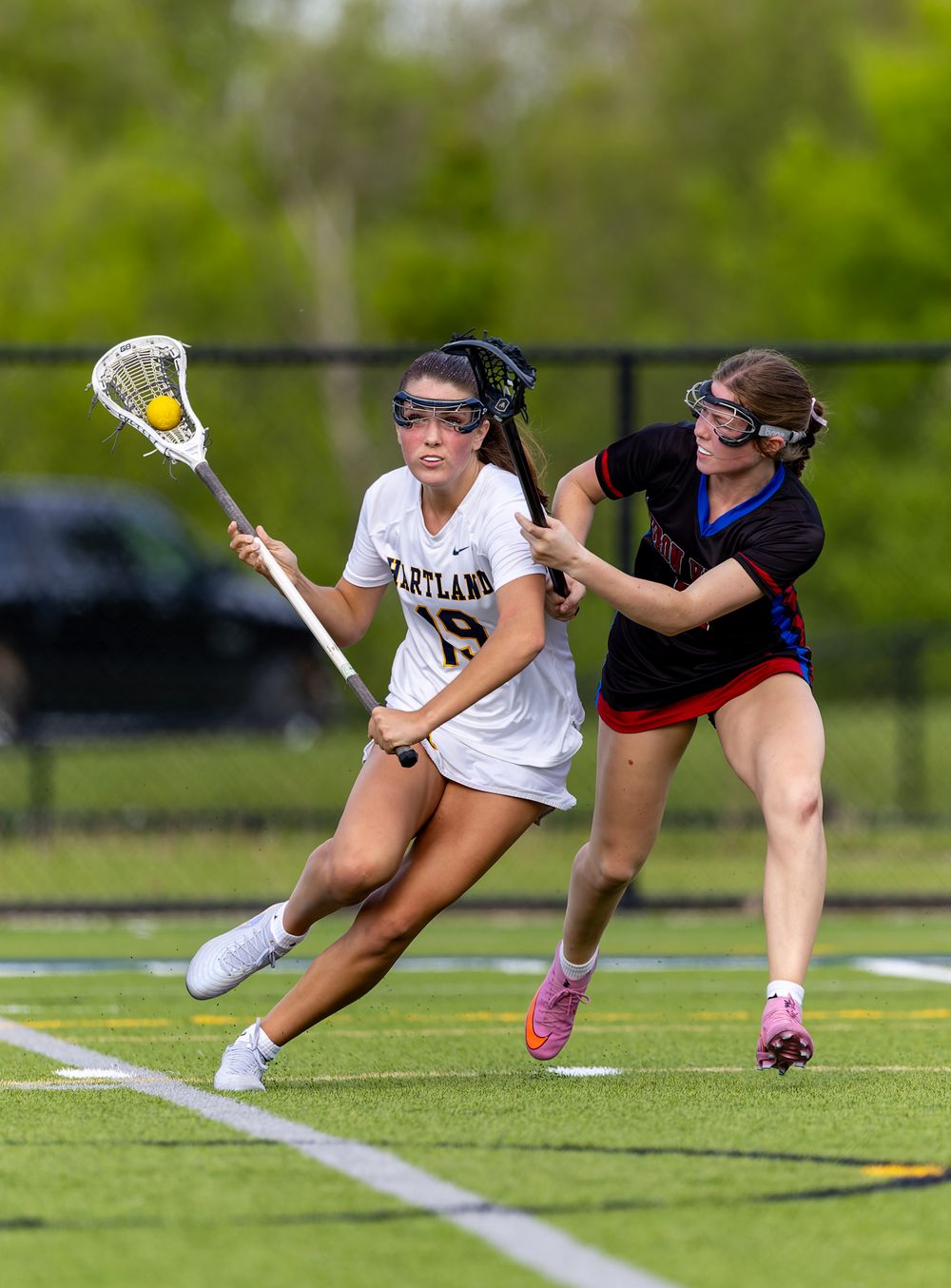 Two female lacrosse players compete on a green turf field, one in white Hartland jersey cradling ball while defender pressures her.