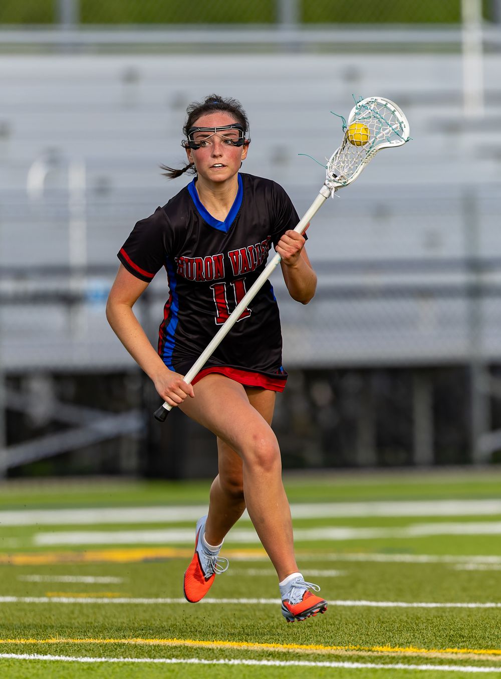 Female lacrosse player in black jersey running with stick and ball during an outdoor game on turf field.