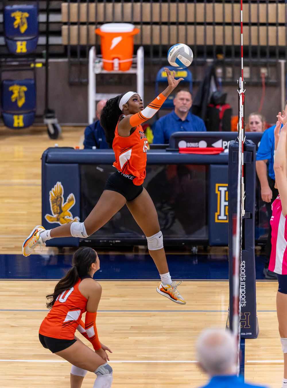 Volleyball player in orange jersey leaps high to spike the ball during an indoor match at a school gymnasium.