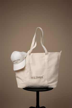White canvas tote bag and baseball cap displayed on black pedestal against brown background.