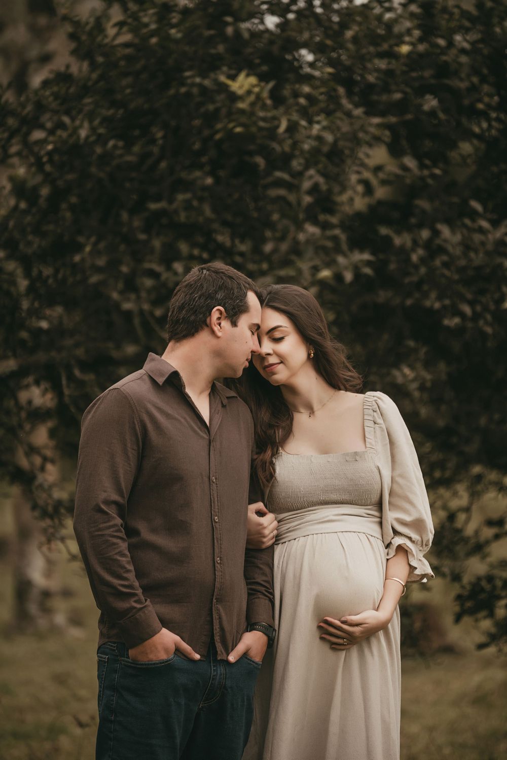 Romantic maternity photo of couple in earthy tones sharing intimate moment outdoors with natural backdrop.