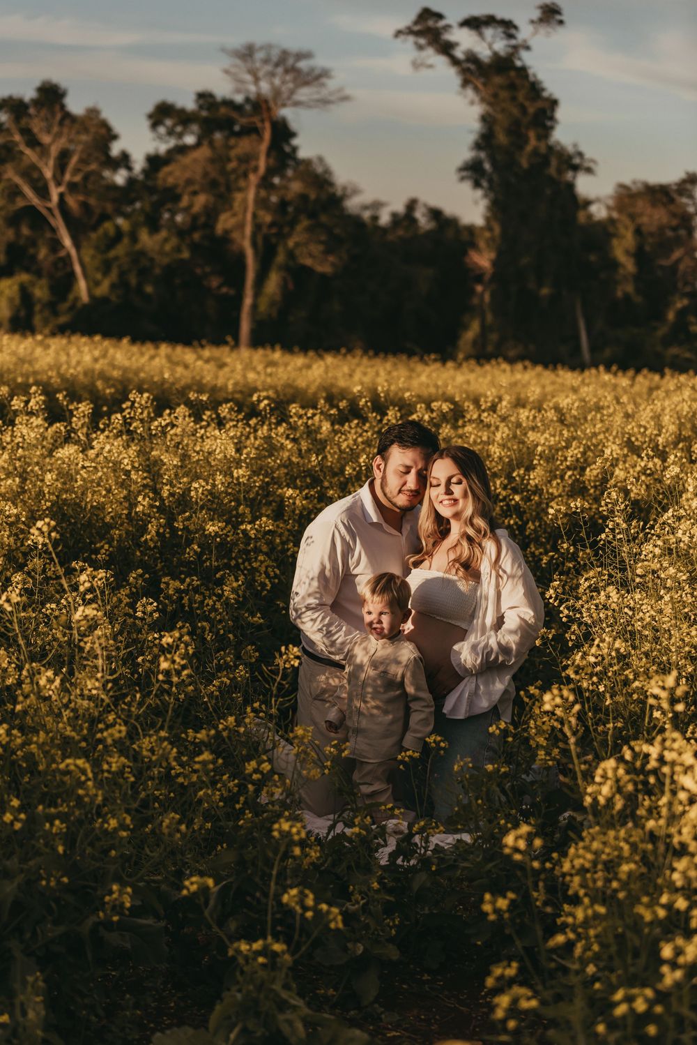 A couple stands together in a golden field of wildflowers at sunset with tall trees in the background.