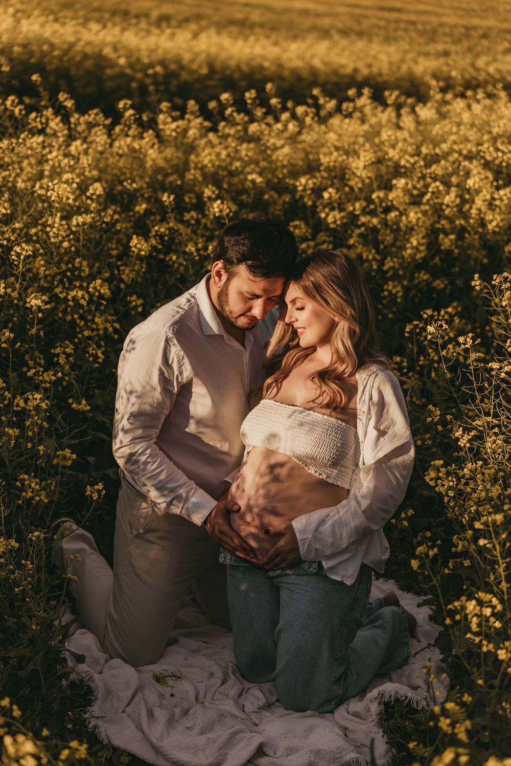 A couple shares an intimate moment while sitting on a blanket in a golden wildflower field at sunset.