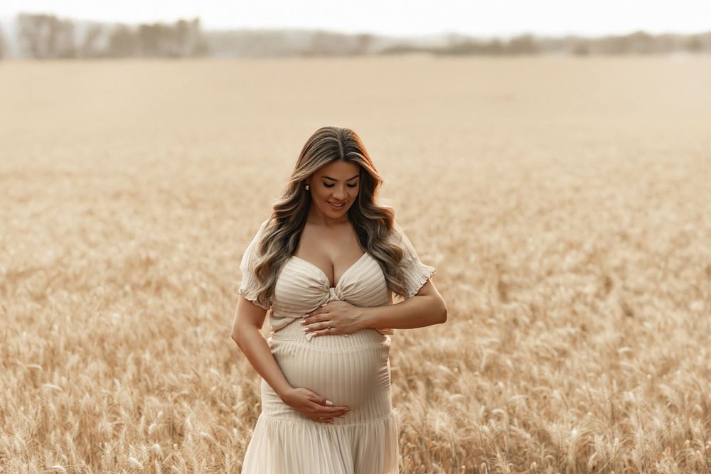 A pregnant woman in a light flowy dress stands serenely in a golden wheat field during sunset.