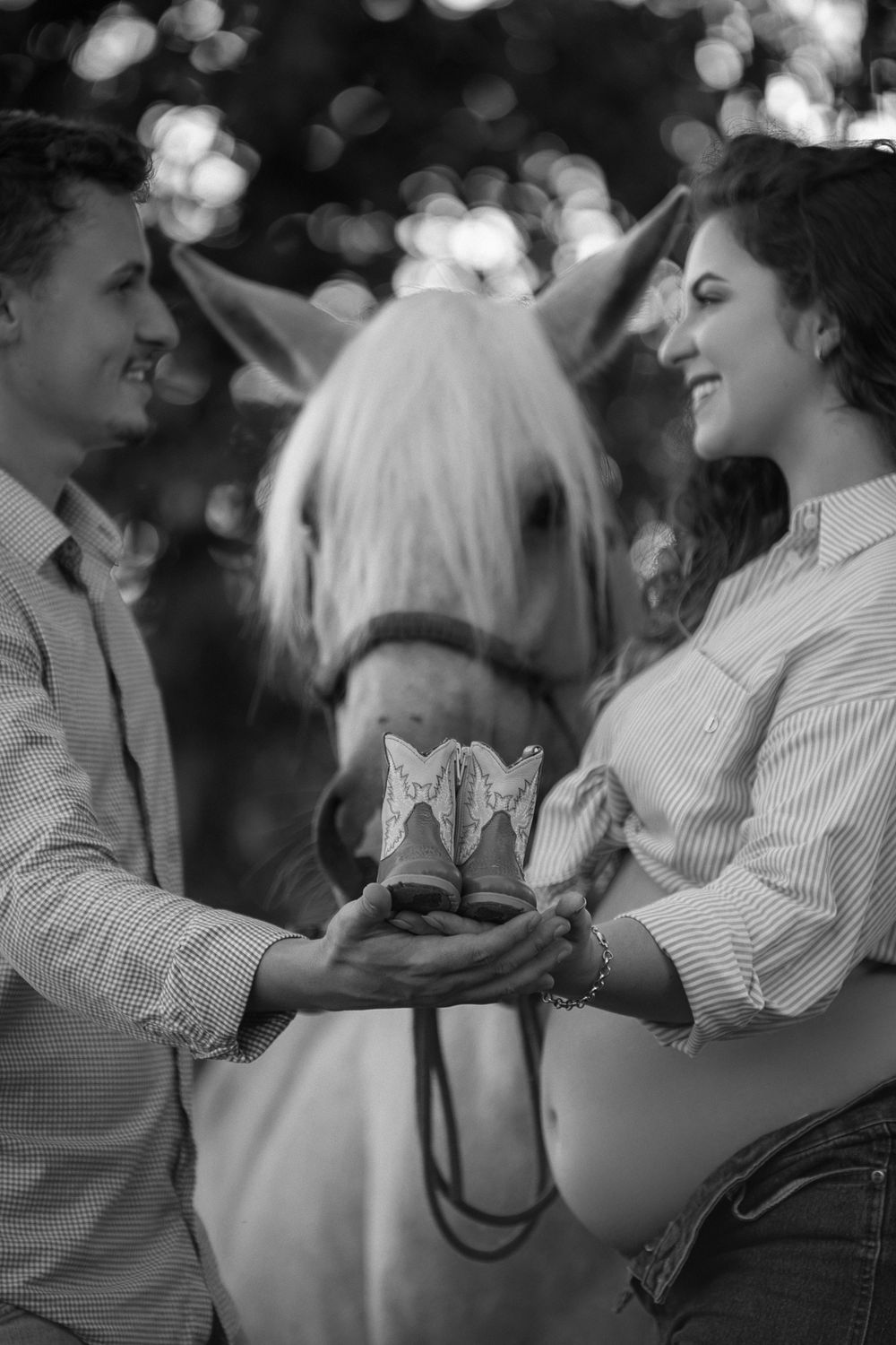 A romantic black and white photo of a couple holding cowboy boots together with a white horse in the background.