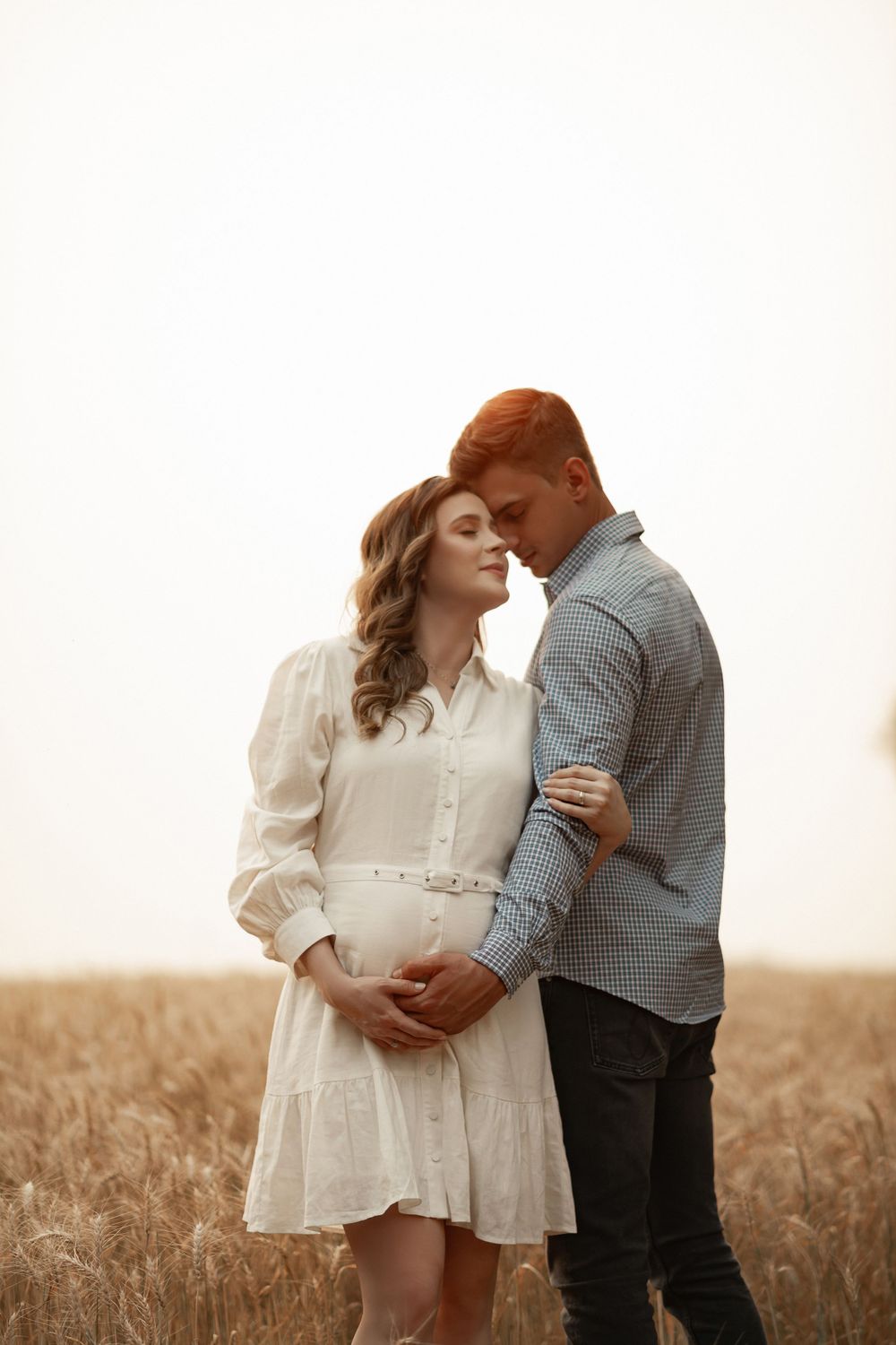 A romantic couple embraces in a golden wheat field at sunset, the woman wearing a flowy white dress.