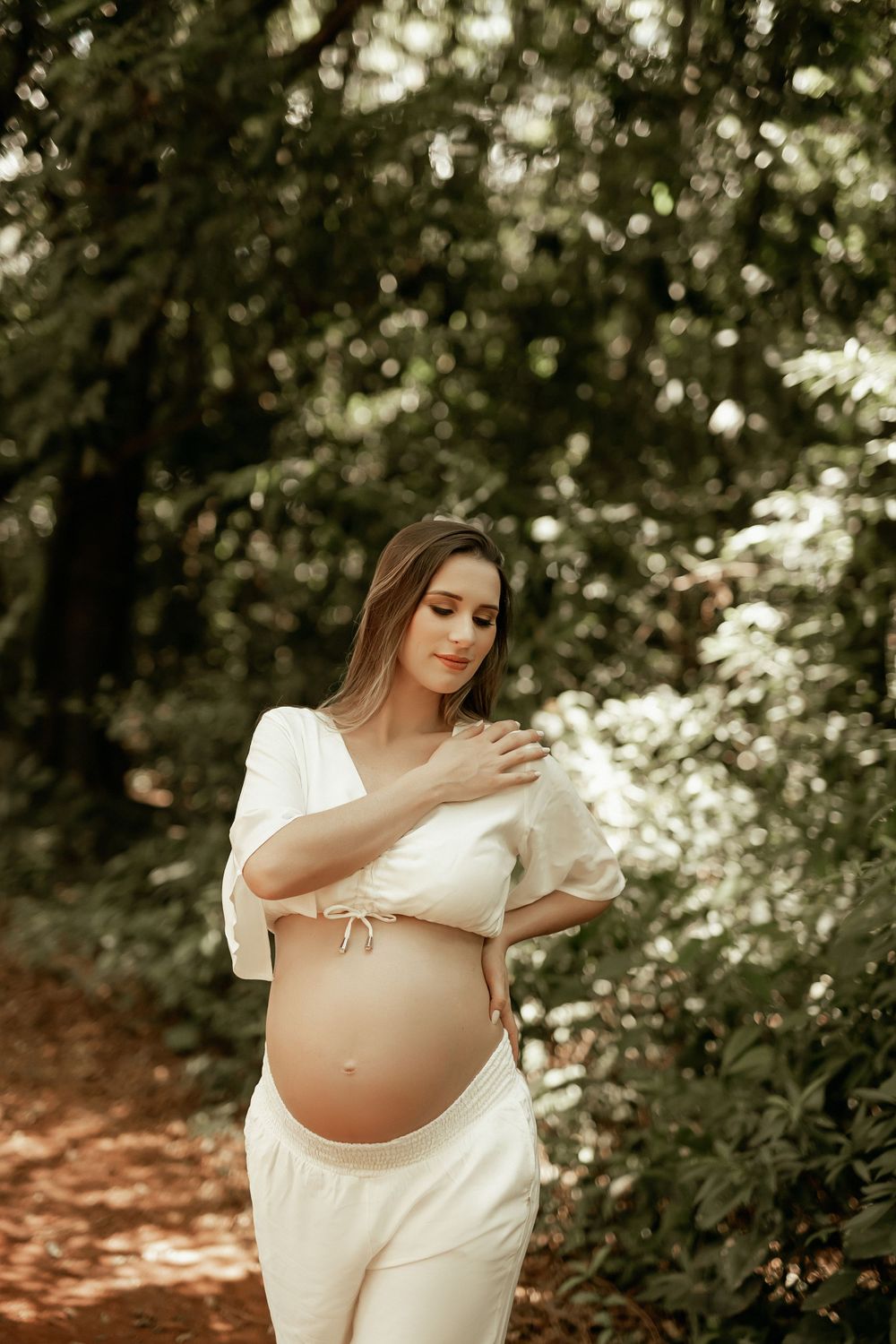 Expectant mother in white dress poses gracefully in a sunlit forest setting during maternity photoshoot.