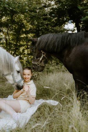 Young girl sitting on a blanket in a field, smiling between a white and dark horse outdoors.