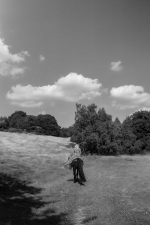 Black and white photo of a person standing in a sunny meadow with trees and clouds in the background.