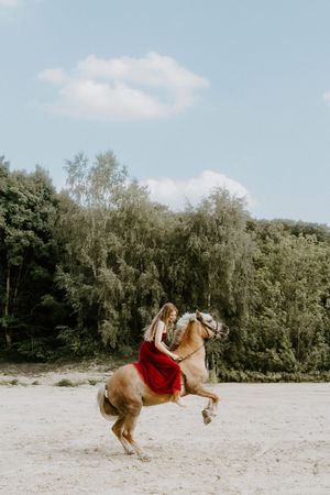 Woman in red dress riding a rearing palomino horse on sandy ground with lush green trees and blue sky backdrop.