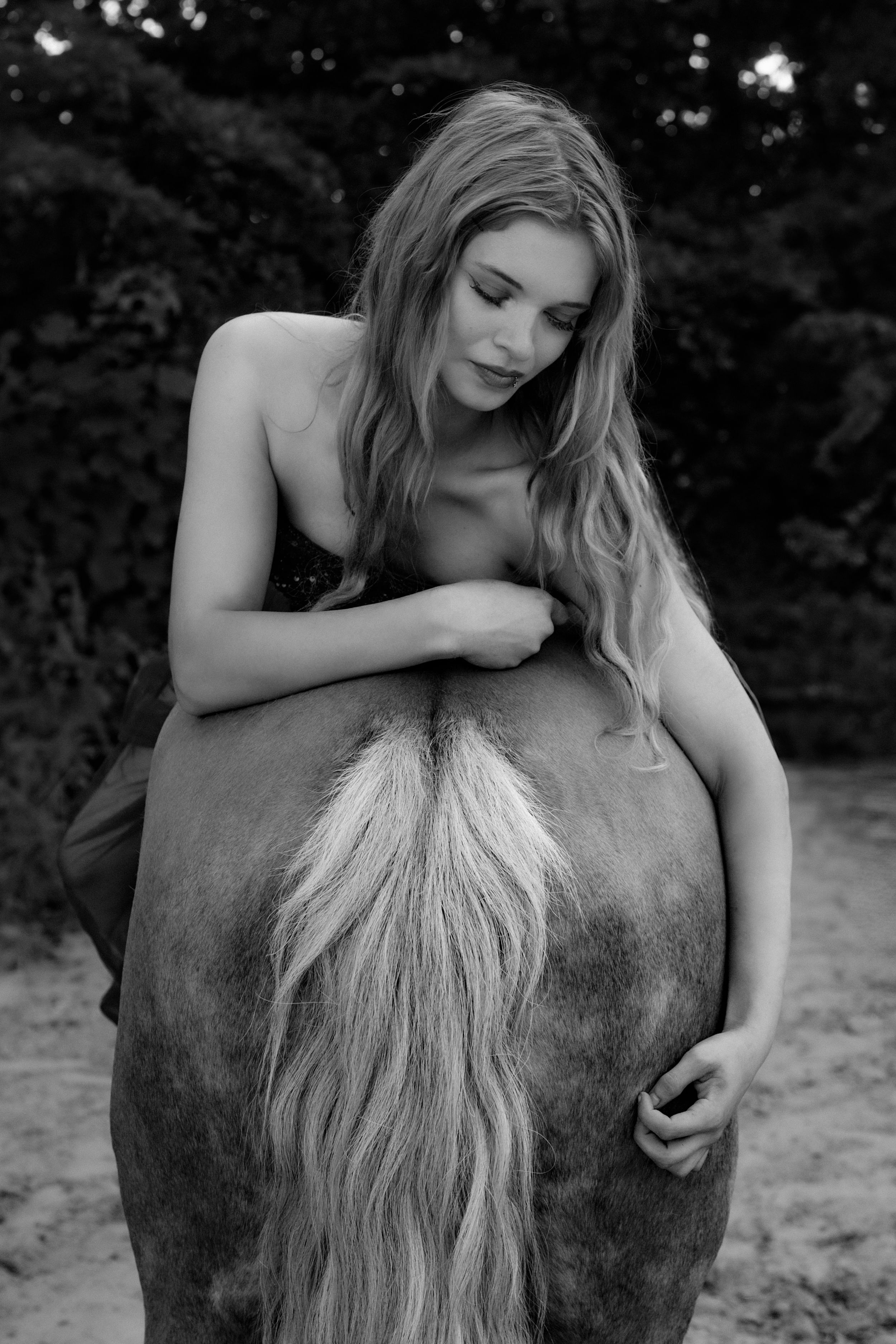 Black and white photo of a young woman with long hair leaning over a horse's back outdoors.