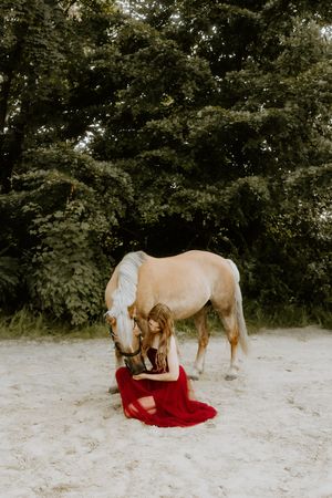 Woman in red dress kneeling beside a palomino horse on sandy ground with lush green trees in background.