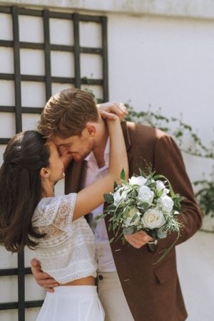 A couple in wedding attire sharing an intimate moment with white flowers against a latticed wall.