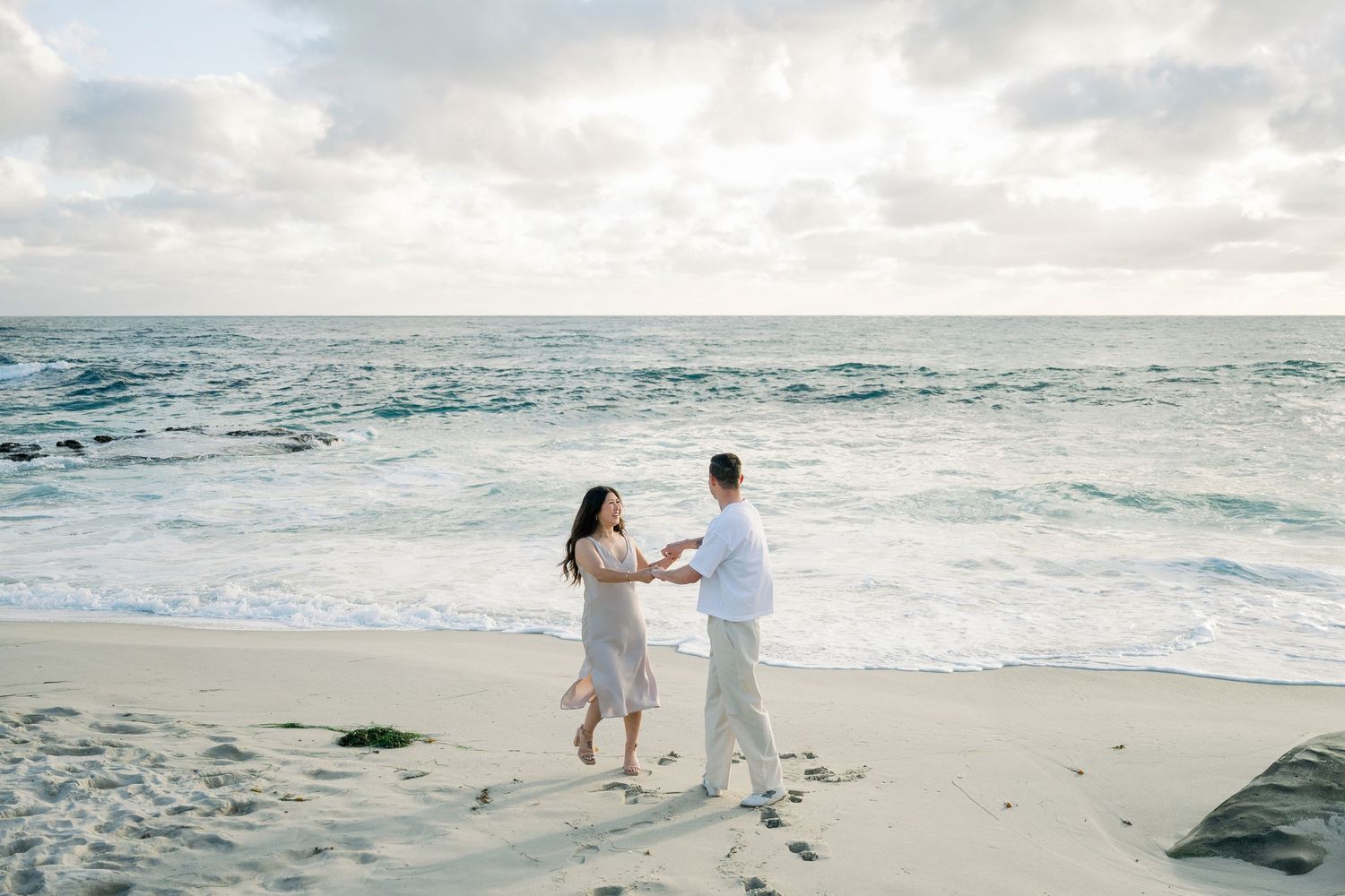 Happy couple walking barefoot on beach in matching light colored formal wear during sunset.