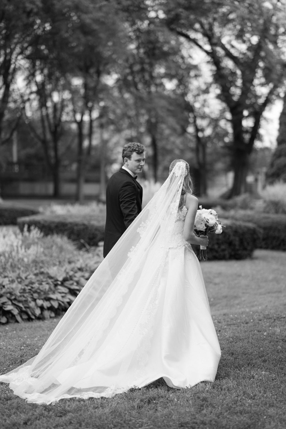 Black and white photo of a bride in a flowing gown and veil holding bouquet, standing with groom in a garden. - The Blackstone Hotel